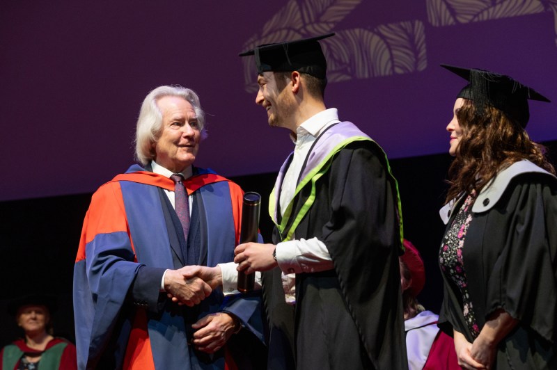 Student wearing cap and gown shaking hands on stage at graduation.