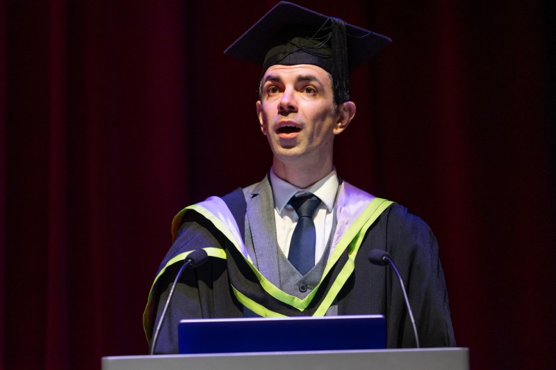 Student wearing cap and gown speaking at Northeastern London graduation ceremony.