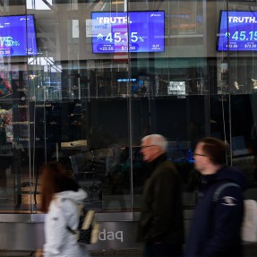 People walking by televisions showing market trading information.