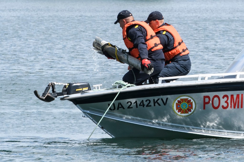 Two people in a boat wearing life jackets remove an unexploded ordnance from a lake.