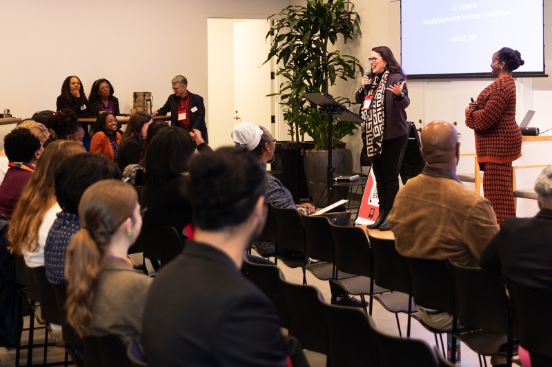 Person speaking at a microphone in a room full of people.