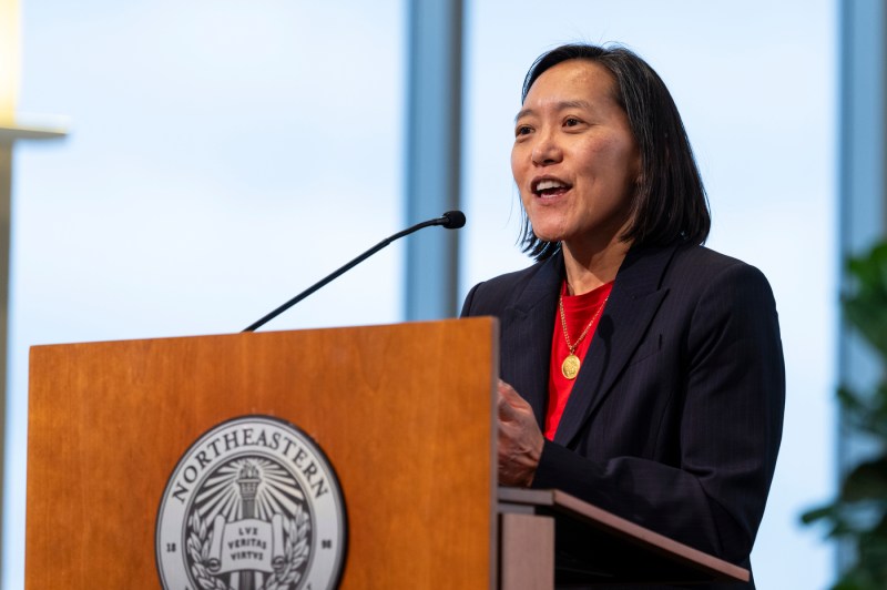 A person wearing a red shirt and black blazer speaking into a microphone at a podium.