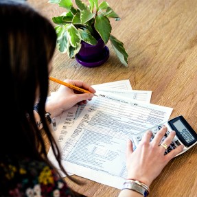 A person filling out paper tax forms using a pencil and a calculator.