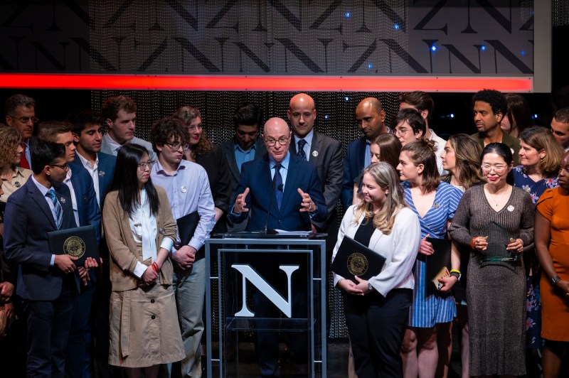 President Aoun standing at a podium in Blackman auditorium speaking while surrounded with a group of people. 