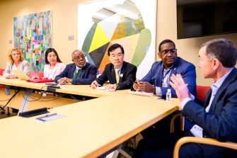 Six people in formal attire sit at a long table in a brightly lit conference room.