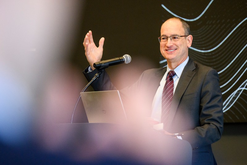 David Luzzi wearing a suit and tie speaking into a microphone and gesturing with his right hand.