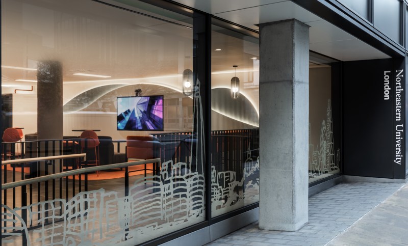 A modern-looking office from the outside, as seen through a large window. The office has orange chairs and a large television.