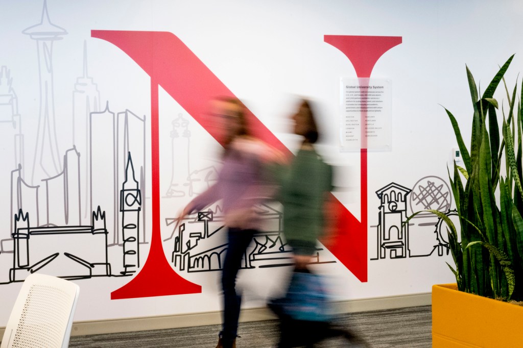 A time lapse photo of two people walking in front of a Northeastern logo at the Portland campus.