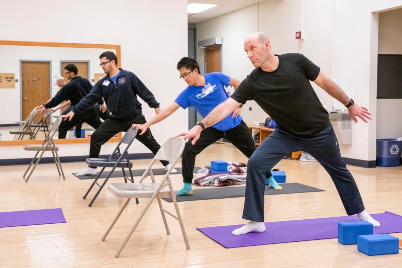 People in a yoga studio, standing on mats, leaning forward with one hand on the back of a chair.