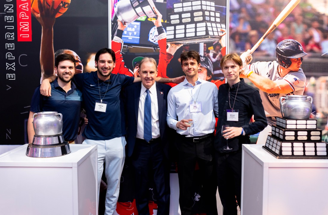 A group of 5 people posing next two two silver trophies.