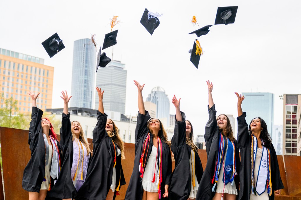 Seven people wearing regalia toss their mortarboards in the air outside on a sunny day.
