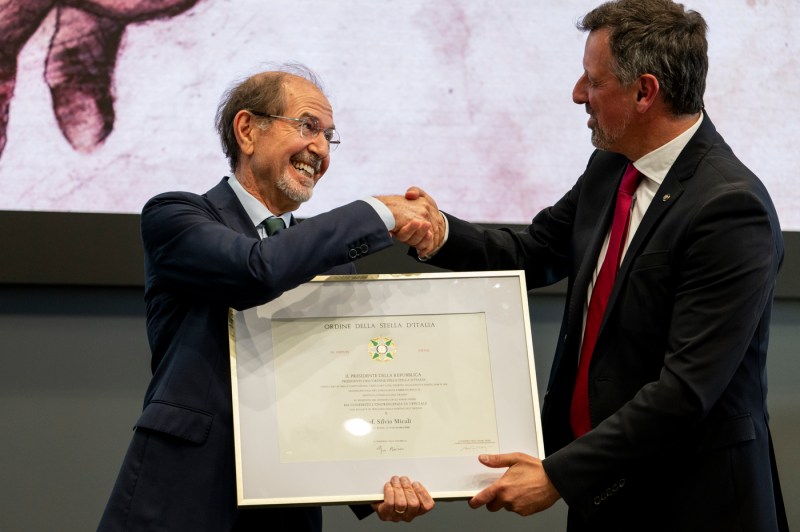 Silvio Micali holding a large award and shaking another person's hand.