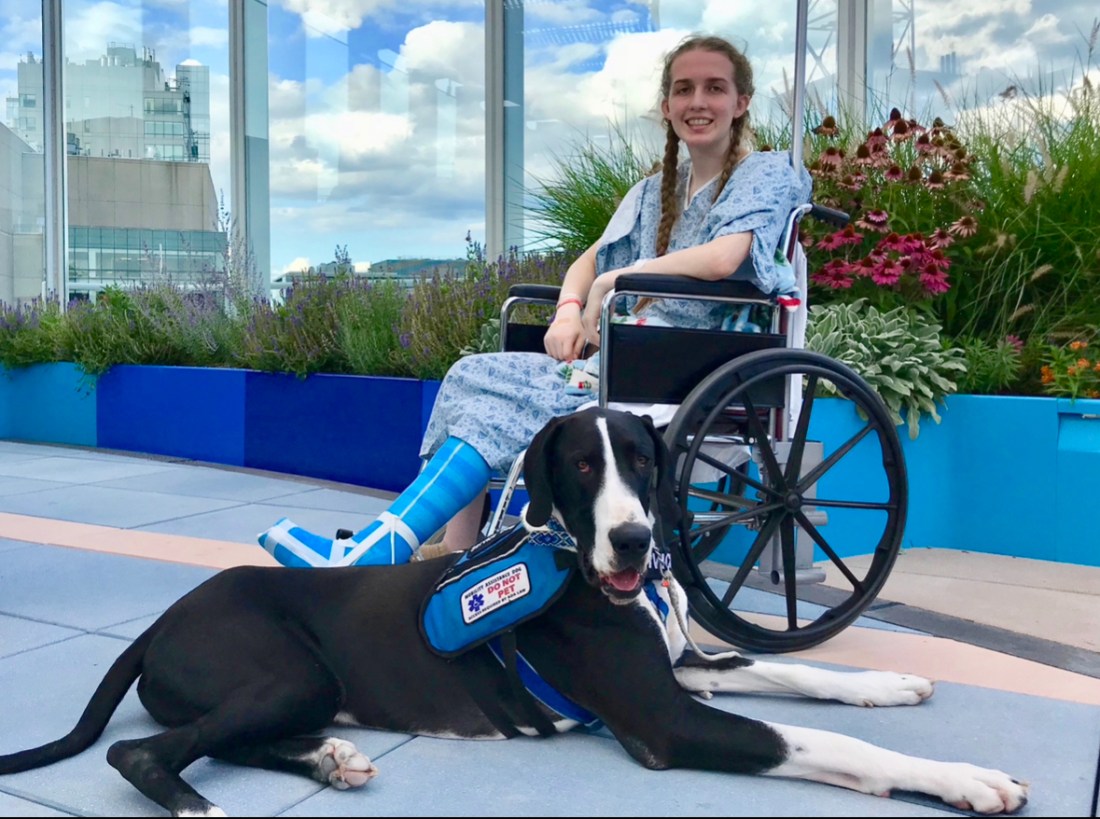 Fiona Howard in a hospital gown sitting in a wheelchair with a black and white dog laying in front of her.