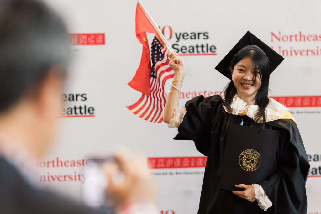 A person wearing regalia is holding two flags while having their picture taken.