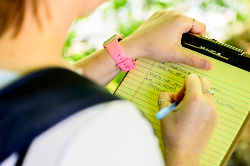 A person takes notes in a notebook while standing outside on a sunny day.