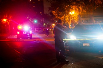 Two police officers stand outside at night near yellow crime tape while a police cruiser's red and blue lights flash in the distance.