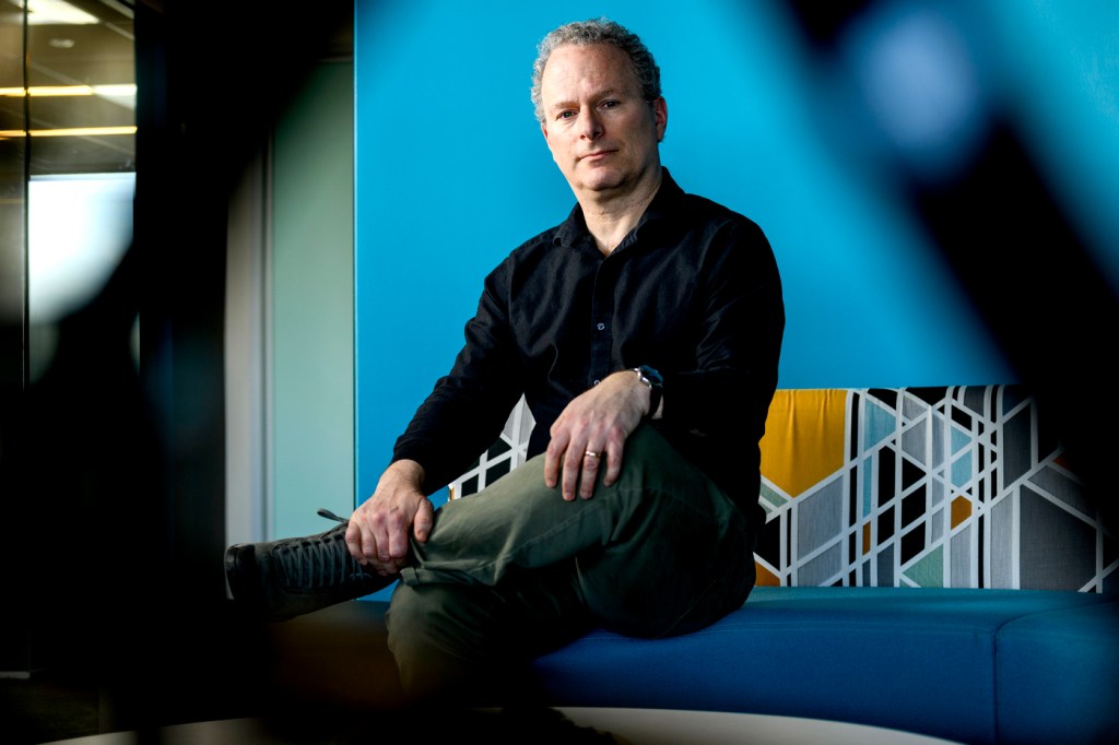 David Lazer sitting in a chair in front of a blue wall with one leg crossed over the other.