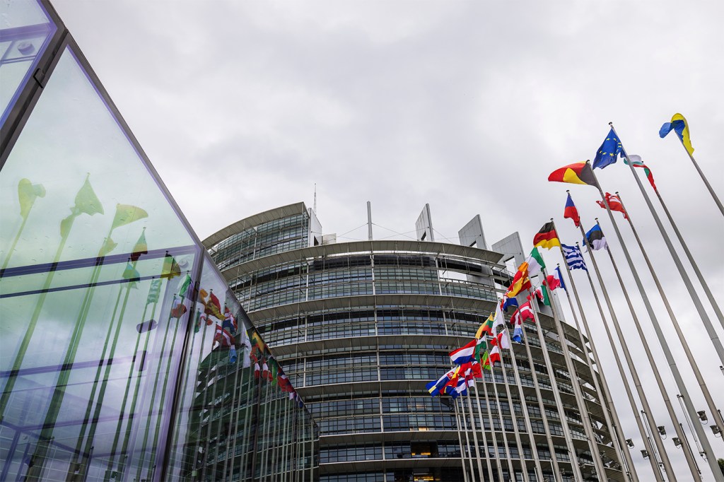 EU flags flying outside of a European Parliament building on a cloudy day.