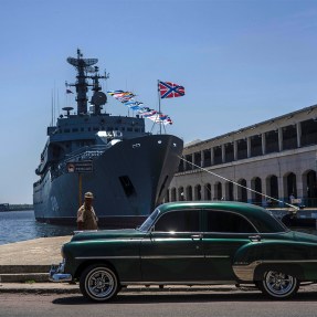 A Cuban sailor walking outside in the sun past a Russian Navy ship.