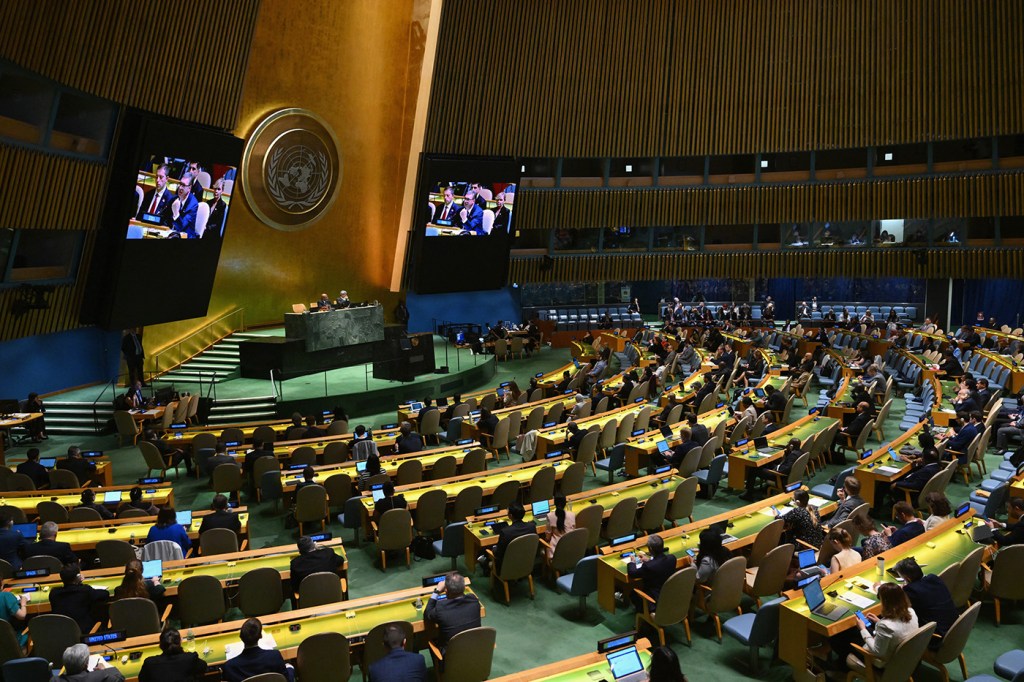 Hundreds of people sit in the United Nations Headquarters, a room with green carpets and wooden benches facing large TV screens.