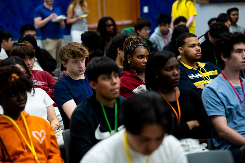 High school students of varying ages, ethnicities, and genders sit in an auditorium watching the presentation.