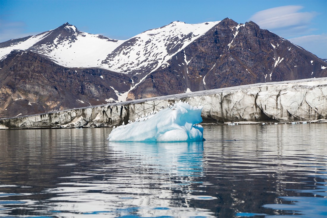 A small iceberg floating in the water in front of snowy mountains.