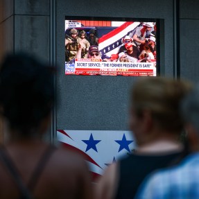 The blurry outline of people is illuminated by a television screen broadcasting political content.