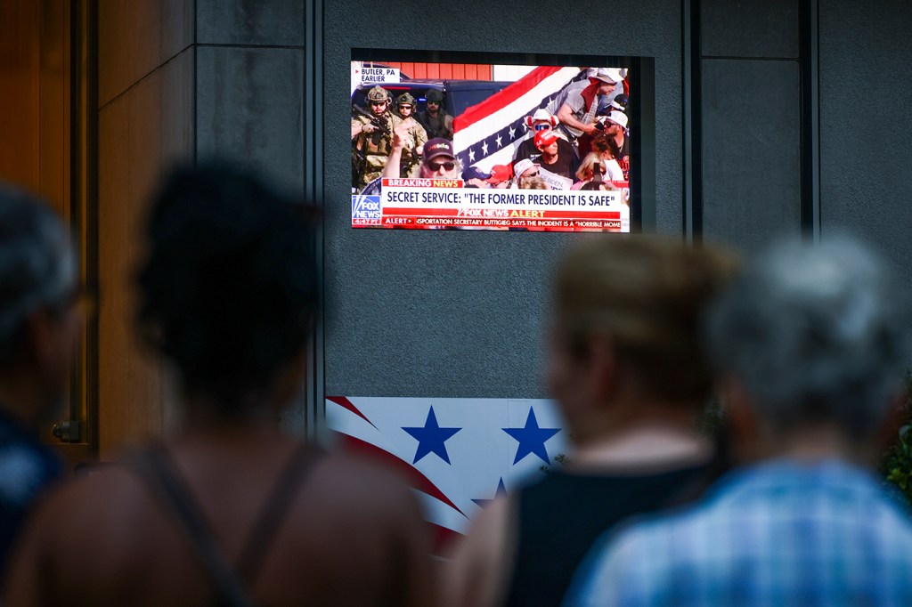 The blurry outline of people is illuminated by a television screen broadcasting political content.