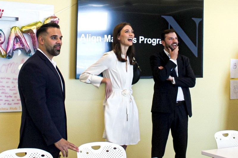 Karlie Kloss wearing a white dress laughing and standing next to two people in suits.