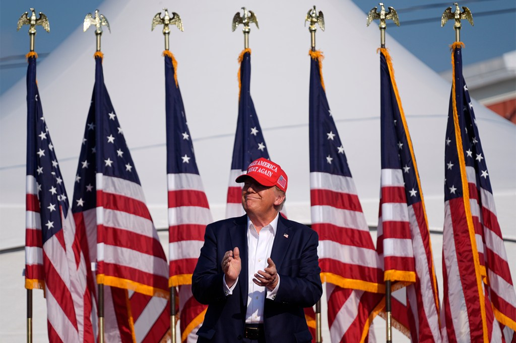 Donald Trump smiles and claps while standing on stage in front of a row of American flags.