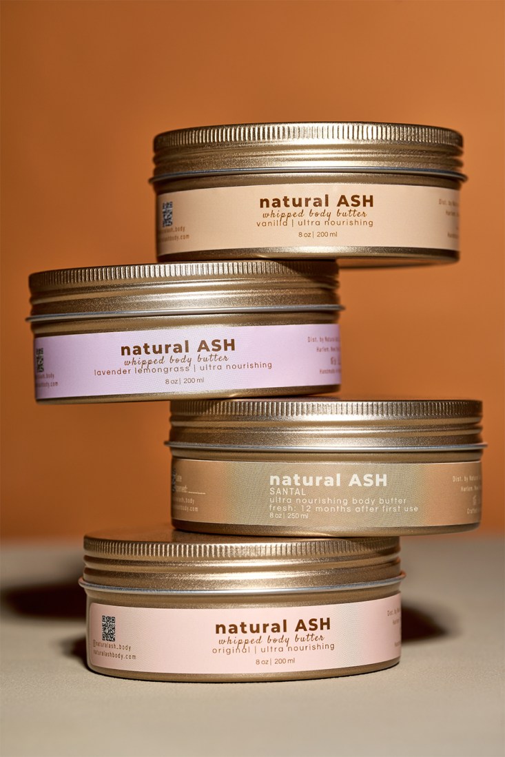 Four Natural Ash body butter containers are stacked unevenly on top of each other, while displaying their labels.