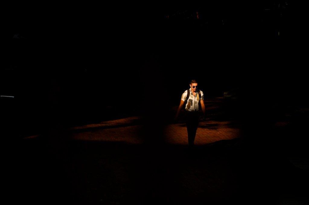 A person wearing a backpack and white button shirt walks out of the shadows and into the sunlight.