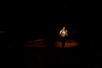 A person wearing a backpack and white button shirt walks out of the shadows and into the sunlight.