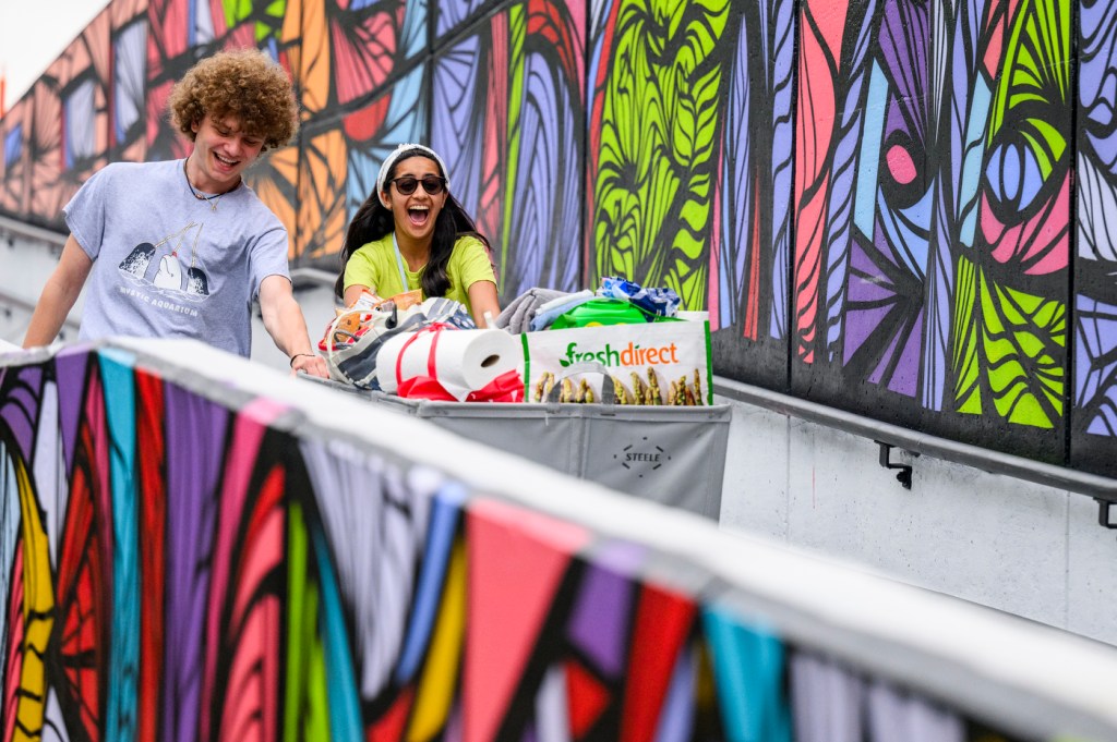 Two people push a bin down a ramp while walking past a multi-colored mural.