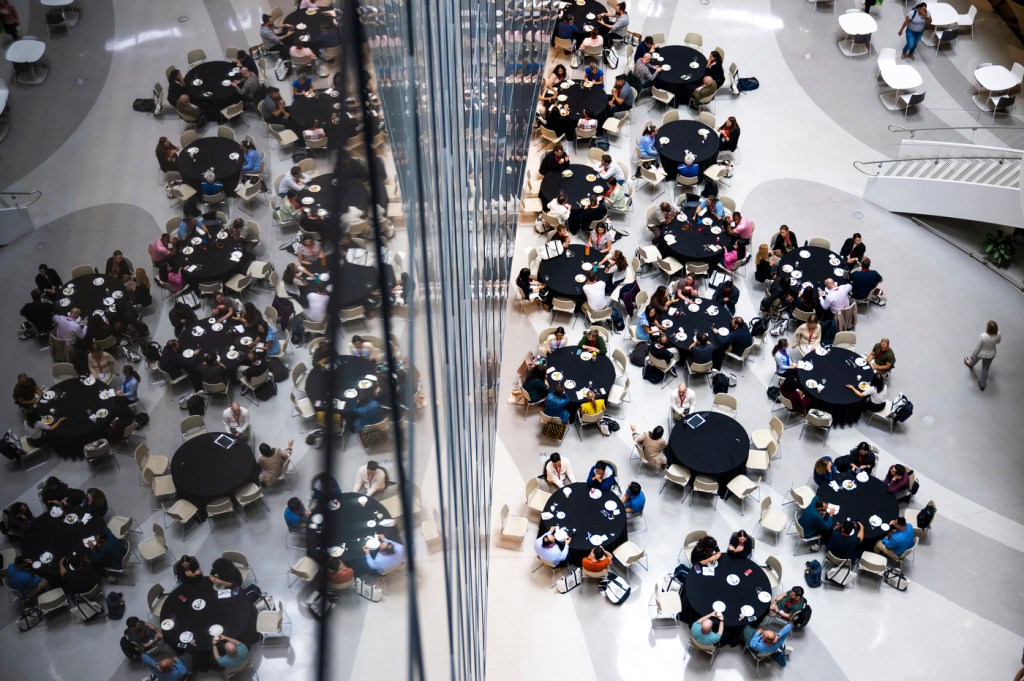 People sit at multiple round, white-colored tables in a brightly lit lobby.