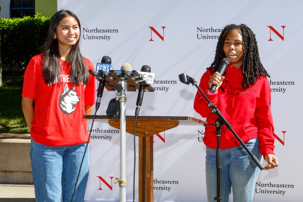 Two students wearing red Northeastern branded sweatshirts speaking into microphones at Oakland Press Conference.