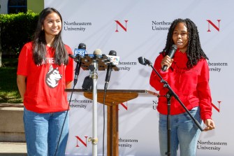 Two students wearing red Northeastern branded sweatshirts speaking into microphones at Oakland Press Conference.