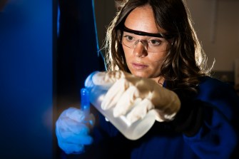 A person wearing a lab coat and goggles pours blue liquid into a test tube.