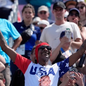 Snoop Dogg wearing a USA shirt at a women's beach volleyball game with his arms raised in the air.