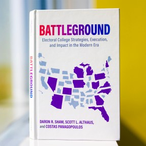 Costas Panagopoulos' new book standing upright on a window sill. The book is white with the word 'Battleground' at the top in a gradient from red to blue. It has a map of the United States with some states shaded in purple.