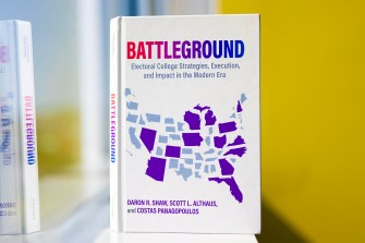 Costas Panagopoulos' new book standing upright on a window sill. The book is white with the word 'Battleground' at the top in a gradient from red to blue. It has a map of the United States with some states shaded in purple.