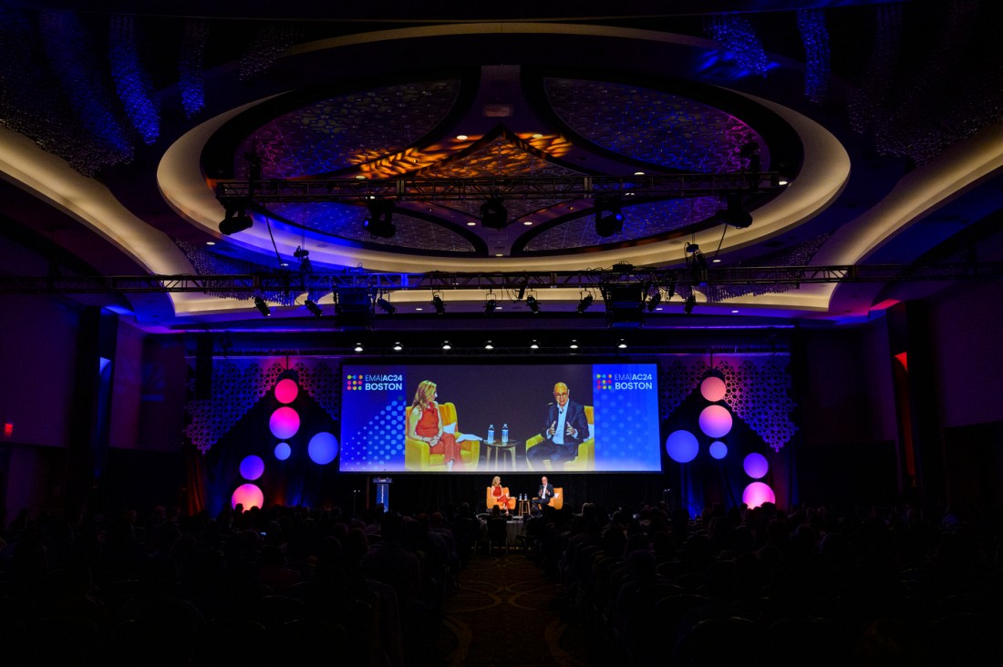 President Aoun and Claire Goldsmith sitting on stage at the Sheraton Boston Hotel in orange chairs. A large screen behind them shows a livestream of them for people in the back of the room.