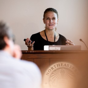 Rebecca Carrier speaking into a microphone at a podium at a Symposium.