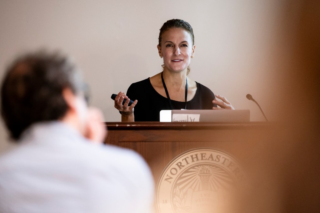 Rebecca Carrier speaking into a microphone at a podium at a Symposium.