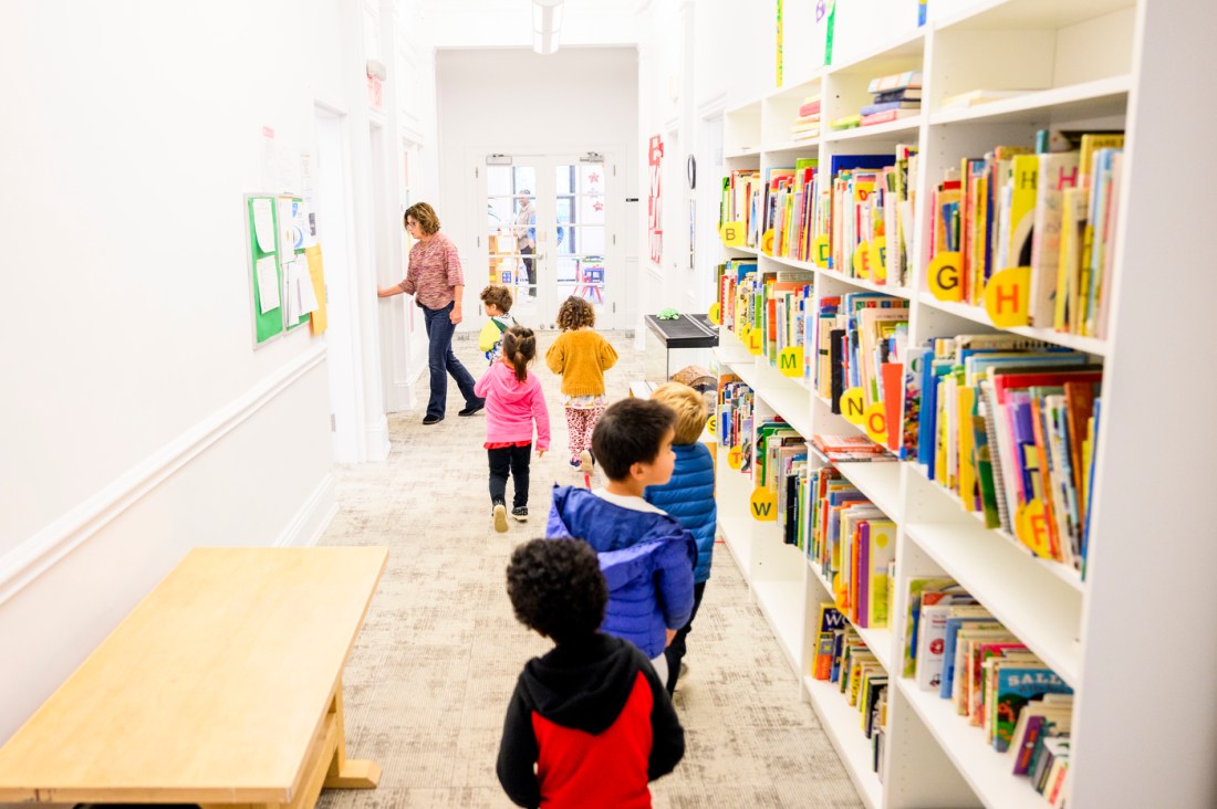 Lee Ann Burdick walking down a hall lined on one side with children's books, with a line of children following her.