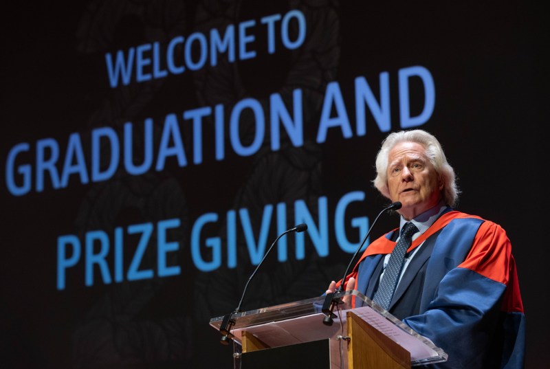 A speaker at the London graduation ceremony standing in front of a black screen that says "Welcome to Graduation and Prize Giving" on it in blue text.