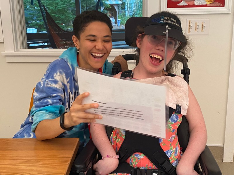 A student holding a laminated sheet of paper in front of a person with a disability. 