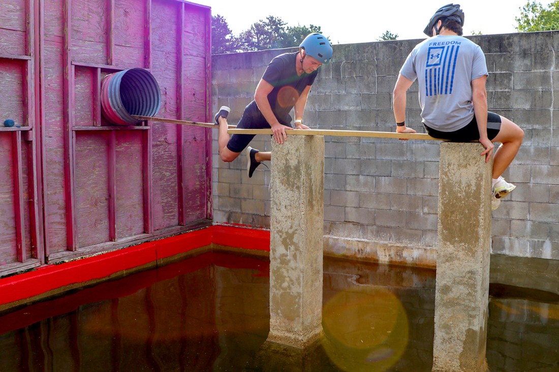 Two students wearing bike helmets balanced on wooden planks balanced on concrete beams sticking out of a pool of water.