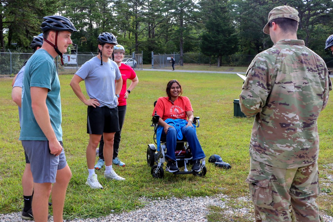 Several students wearing bike helmets, one sitting in a wheelchair, in front of person in a military uniform reading off of a piece of paper.