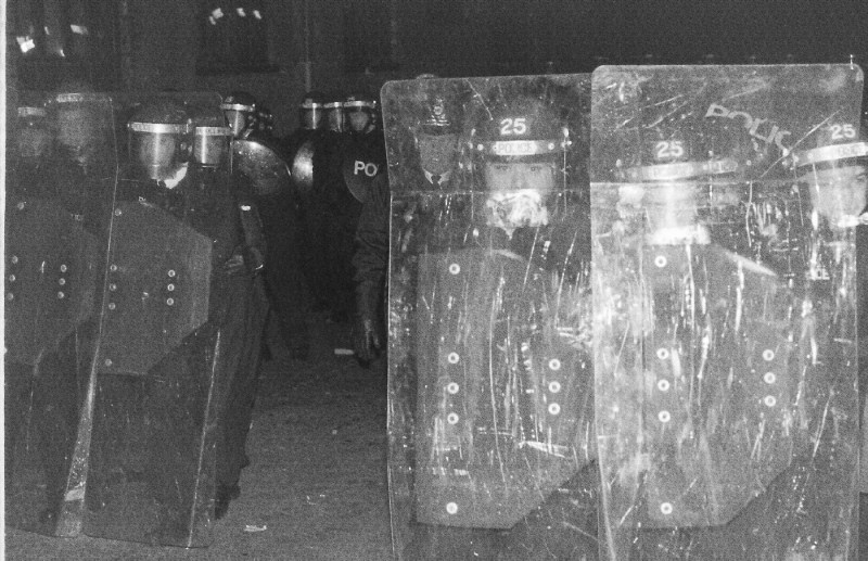 A black and white photo of police officers in riot gear at night.